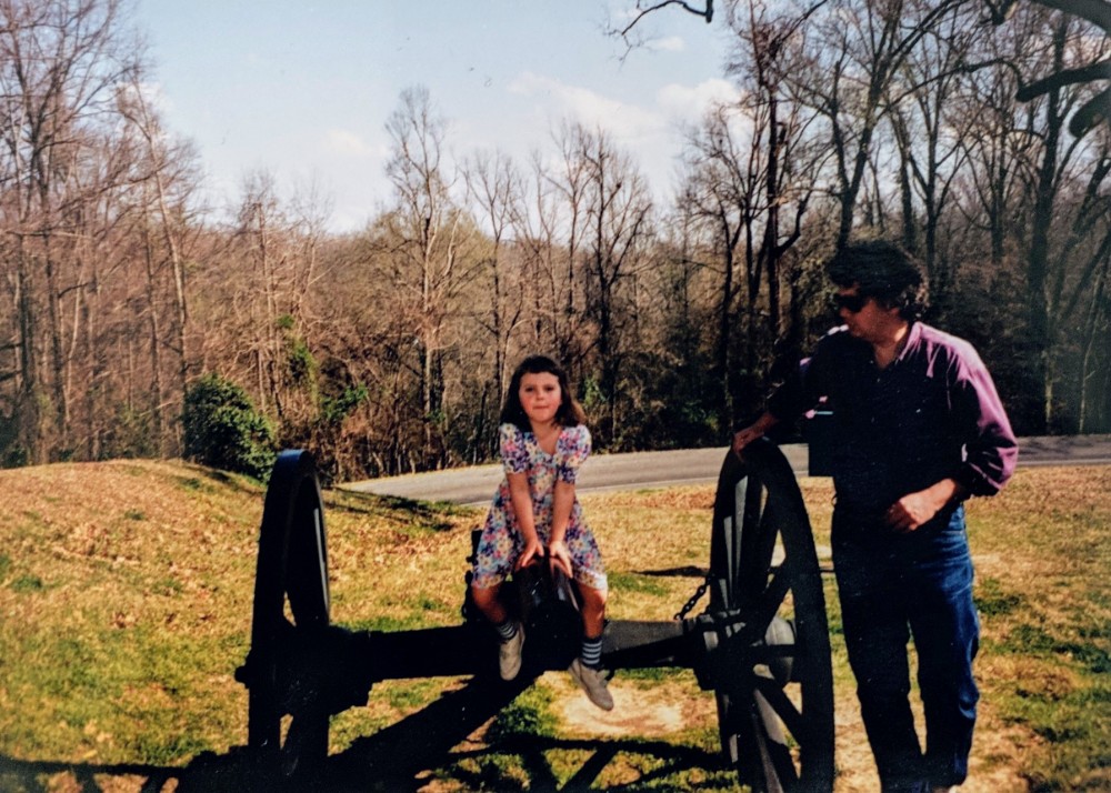 A young girl in a floral dress sitting on a cannon with her dad standing beside her.